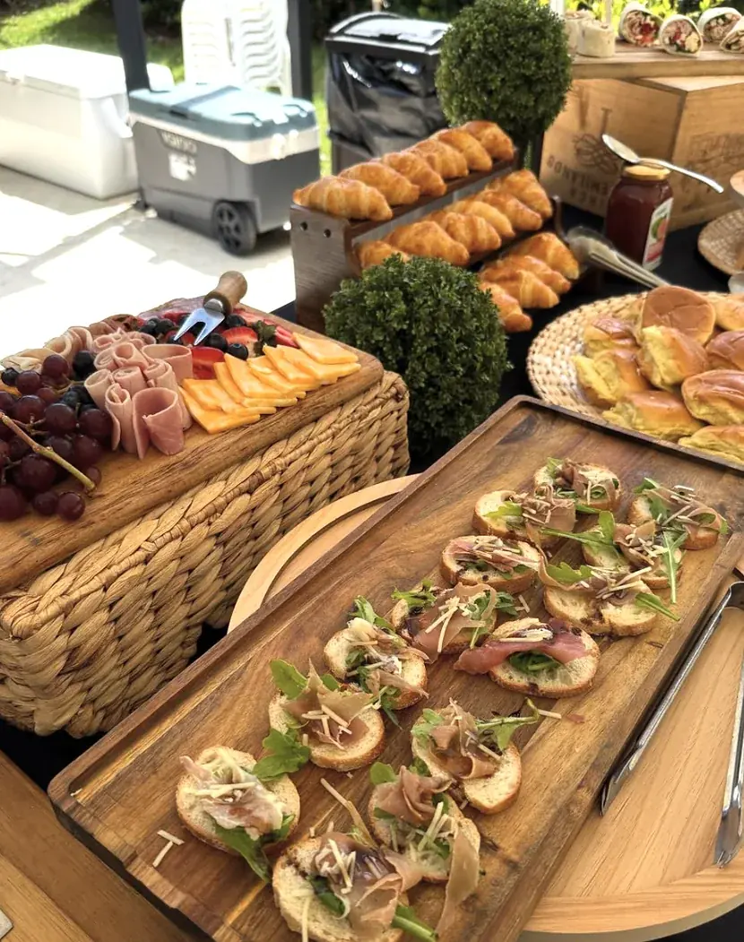 Montaje de brunch con croissants, tabla de jamón, quesos y frutas, y crostini de prosciutto con arúgula