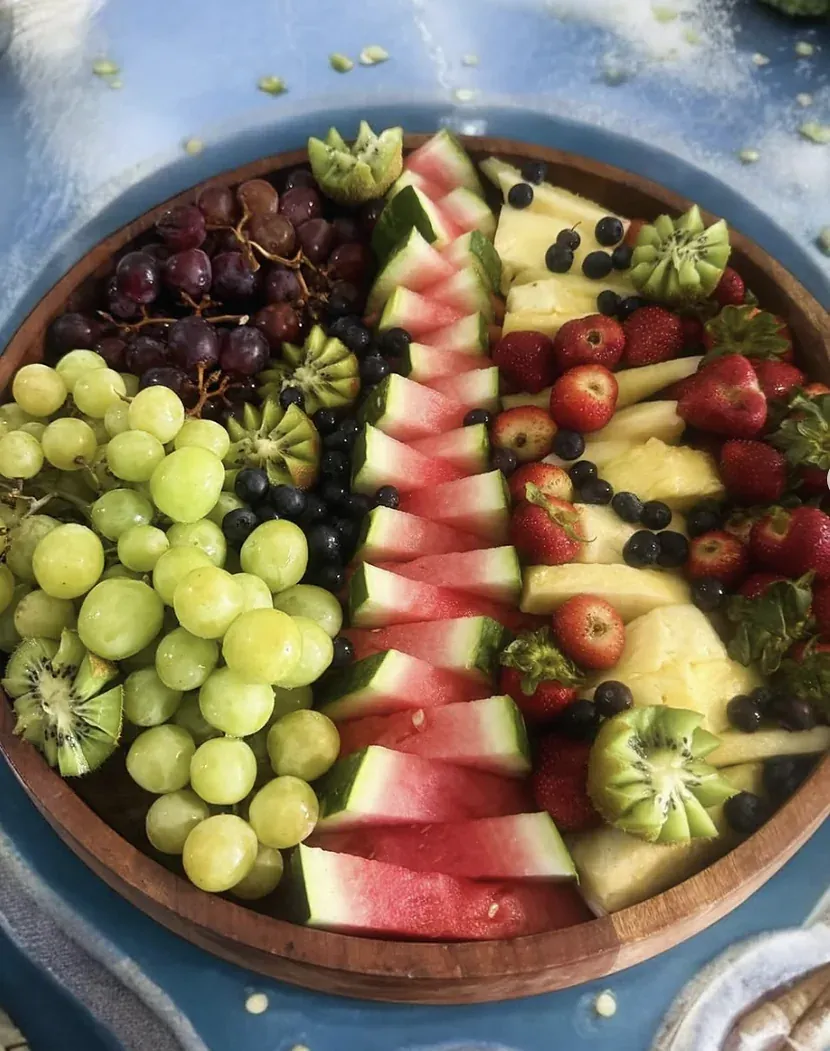 Bandeja redonda de frutas cortadas en patrón: sandía, uvas verdes y moradas, piña, fresas, kiwi y arándanos