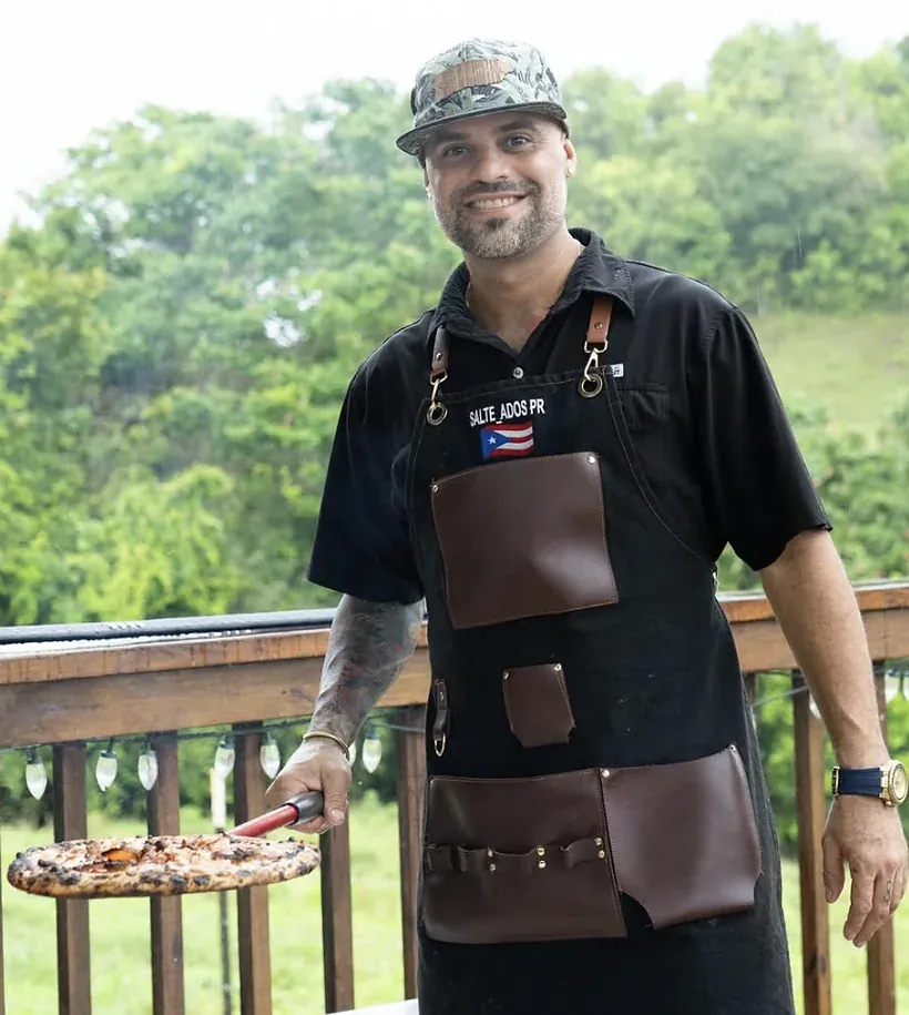 El chef de SalteadosPR con su delantal de cuero de la marca y el parcho de la bandera de Puerto Rico, sosteniendo una pizza recién salida del horno