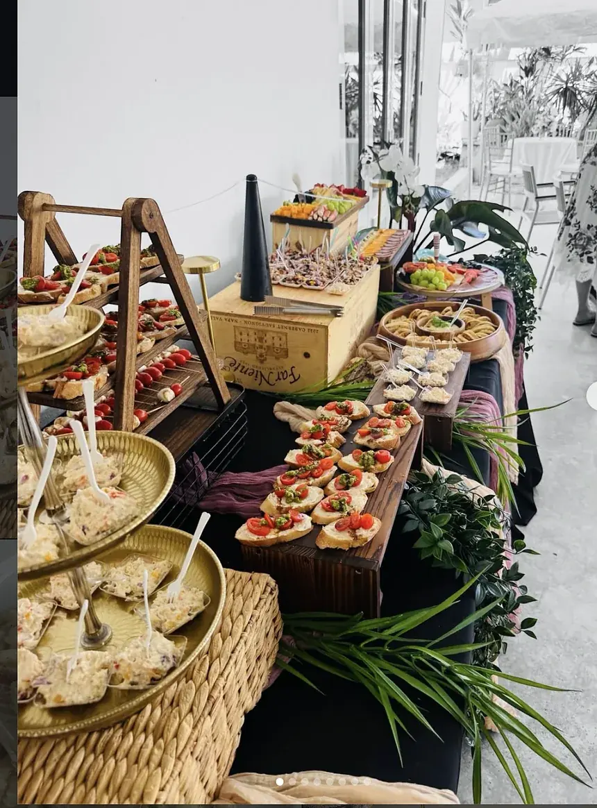 Mesa larga de aperitivos con crostini de tomate, ensaladillas en bandejas doradas, brochetas y greenery, montada para un evento