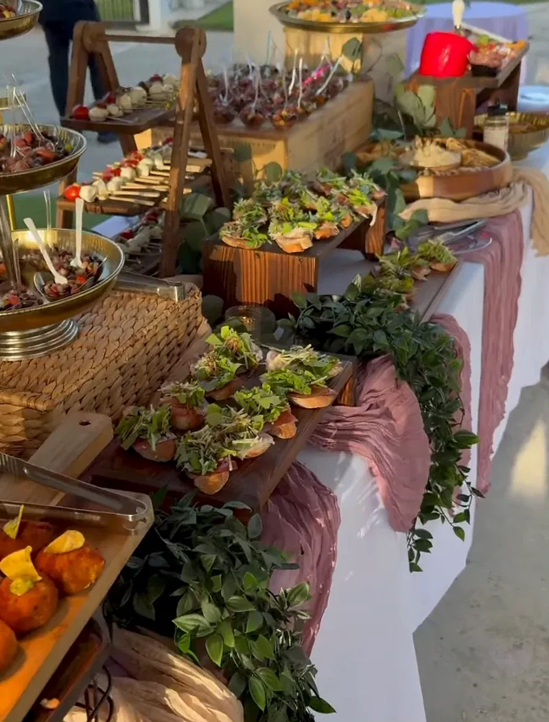 Montaje de boda al atardecer: mesa larga con mantel blanco, caminos de tela rosa viejo, cajas de madera con crostini, quesos y greenery