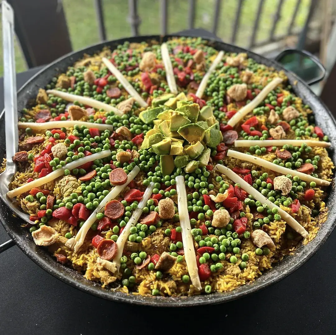 Paella grande decorada en patrón radial, con espárragos en forma de rayos, aguacate picado en el centro, guisantes, pimientos y chorizo