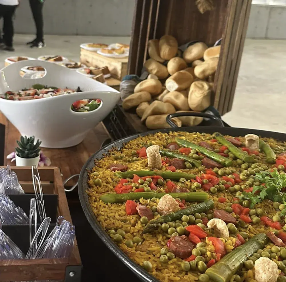Paella con espárragos, guisantes, pimientos rojos y chorizo, junto a una canasta de panecillos y ensaladas en un evento