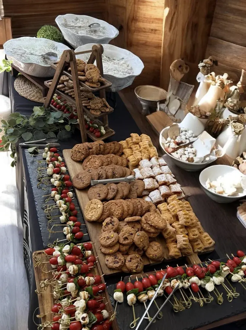 Mesa de postres con galletas, merengues, alfajores cubiertos de azúcar glass y brochetas de tomate y mozzarella
